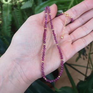 Può includere: Un delicato set di collana e braccialetto con pietre preziose rosa intenso sfaccettate, intervallate da piccole perline color oro. La collana è tenuta in una mano su uno sfondo di fogliame verde.