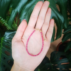 Peut inclure: Un bracelet délicat en pierres précieuses roses avec un fermoir en or. Le bracelet est composé de petites perles rondes et est présenté sur une main sur un fond de feuilles vertes.