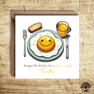 May include: A white card with a cartoon illustration of a fried egg with a smiley face yolk. The card has the text "Happy Birthday to a good egg! Timothy" on it.