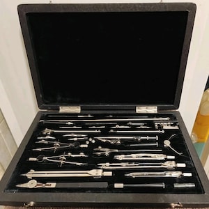 May include: An open, black wooden case with a black velvet interior, filled with various silver drafting tools. The tools include compasses, dividers, and other precision instruments, neatly arranged. The case is likely used for technical drawing or engineering purposes.
