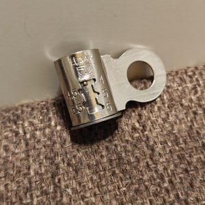May include: A silver crimp connector with a circular eyelet and a cylindrical barrel. The barrel is marked with "34A", "4.5", "5", "5.5", and "6". This connector is used for electrical wiring.