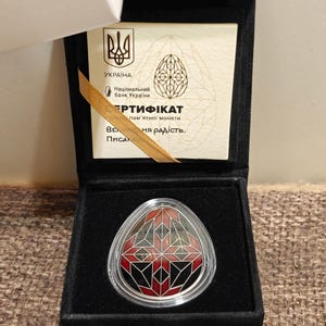 May include: A commemorative coin in a black velvet box. The coin has a red and black geometric design within a clear border. The box contains a certificate with Ukrainian text and a wooden accent.