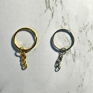 May include: Two keychains, one gold-toned and one silver-toned, are displayed against a marbled background. Each keychain features a circular ring with a small chain of three links attached, designed for holding keys or other small items.