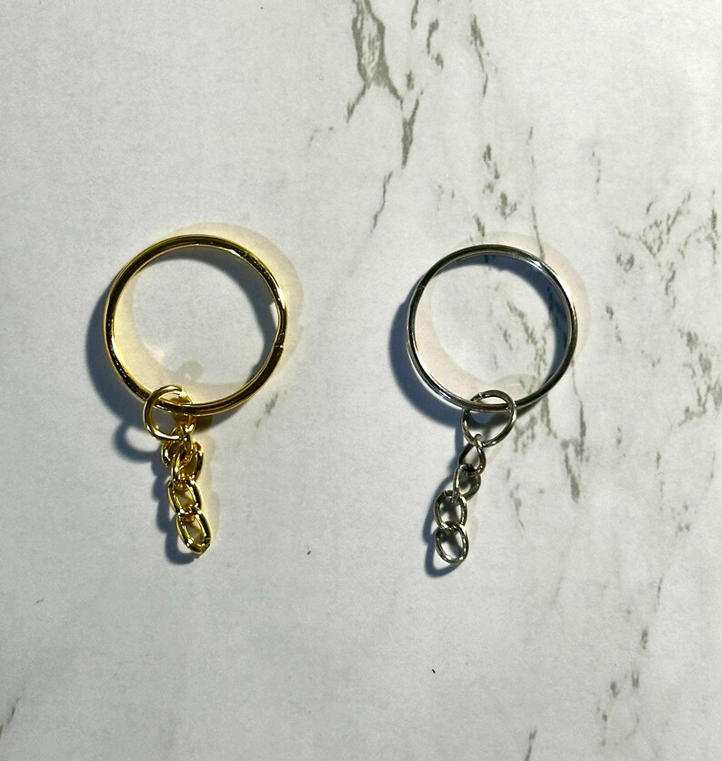 May include: Two keychains, one gold-toned and one silver-toned, are displayed against a marbled background. Each keychain features a circular ring with a small chain of three links attached, designed for holding keys or other small items.