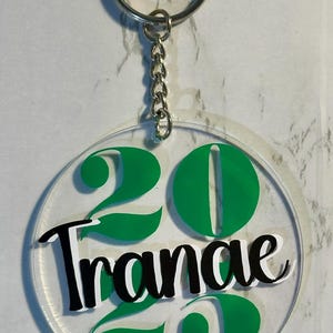 May include: Clear acrylic keychain with a silver chain and ring. The keychain features the number "20" in green, with the word "Trance" in black and white script. The design is circular.