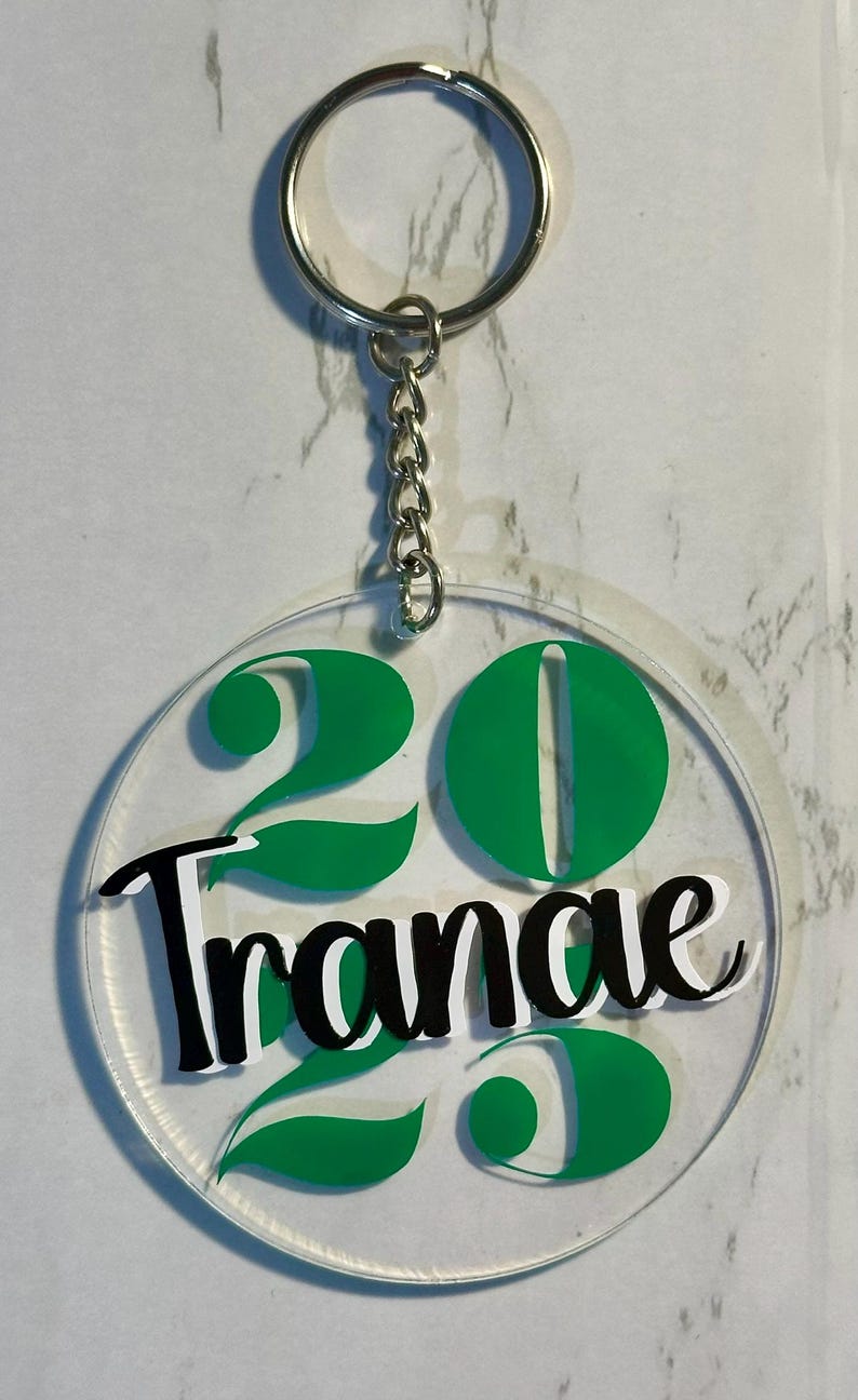 May include: Clear acrylic keychain with a silver chain and ring. The keychain features the number "20" in green, with the word "Trance" in black and white script. The design is circular.