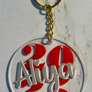 May include: Clear acrylic keychain with a gold-colored ring and chain. The keychain features the name "Aliya" in silver lettering with a white outline, and the number "23" in red. A personalized accessory.