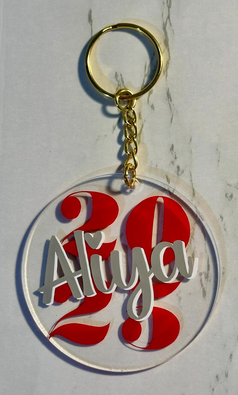 May include: Clear acrylic keychain with a gold-colored ring and chain. The keychain features the name "Aliya" in silver lettering with a white outline, and the number "23" in red. A personalized accessory.