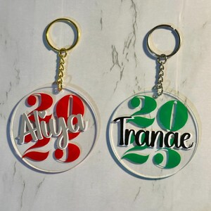 May include: Two clear acrylic keychains with gold-colored key rings. One keychain has the name "Aliya" in silver and red, with the number "23". The other keychain has the name "Tranae" in black and green, with the number "23".
