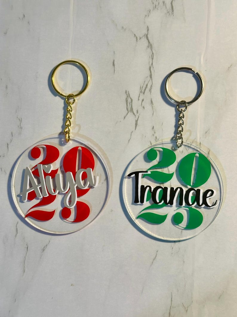 May include: Two clear acrylic keychains with gold-colored key rings. One keychain has the name "Aliya" in silver and red, with the number "23". The other keychain has the name "Tranae" in black and green, with the number "23".