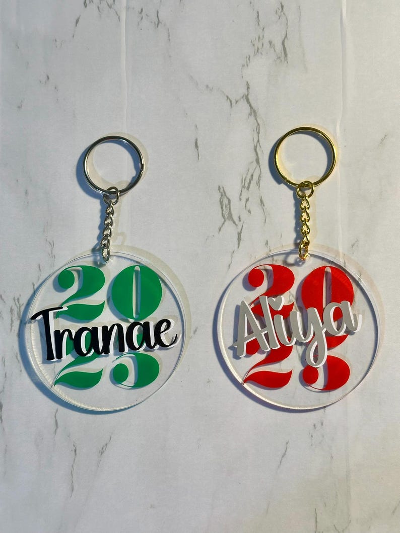 May include: Two clear acrylic keychains with silver and gold key rings. One keychain has the name "Tranae" in black with green accents and the number "22" in green. The other keychain has the name "Aliya" in silver with red accents and the number "22" in red.