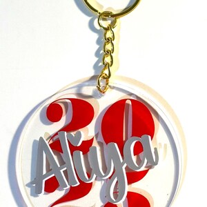 May include: A clear acrylic keychain with a gold-colored ring and chain. The keychain features the name "Aliya" in gray lettering, with the number "23" in red. The design is circular.