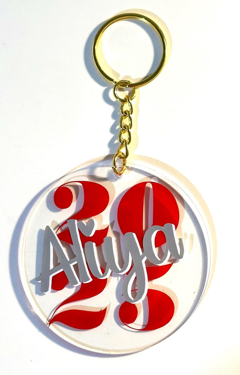 May include: A clear acrylic keychain with a gold-colored ring and chain. The keychain features the name "Aliya" in gray lettering, with the number "23" in red. The design is circular.