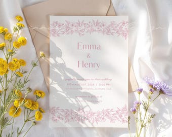 Op de afbeelding: Een trouwkaart met de namen "Emma & Henry" in roze. De kaart heeft een bloemenrand en bevat de datum, tijd en locatie van de bruiloft. Een envelop en bloemen zijn op de achtergrond.