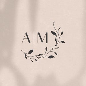 Może przedstawiać: Logo z monogramem z literami "A|M" otoczonymi stylizowanym wieńcem kwiatowym. Wieniec składa się z delikatnych gałązek i liści.