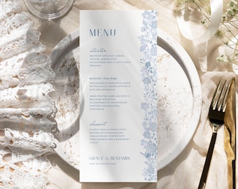 May include: A wedding menu card with a floral design in light blue. The menu lists starter, main course, and dessert options. The card is on a white plate with a gold fork and knife.