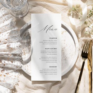 Pode incluir: Um cartão de menu formal com a palavra "Menu" em escrita elegante. O menu lista uma entrada de beterraba e queijo de cabra, um prato principal de salmão grelhado e uma sobremesa de panna cotta de baunilha. O cartão está em um prato branco.