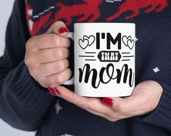 Peut inclure: Mug en céramique blanche avec le texte "I'M THAT mom" en script noir. La tasse présente des motifs de cœurs et de lignes. La tasse est tenue par une personne portant un pull bleu marine.