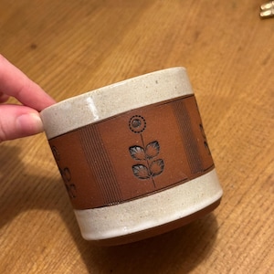 May include: A ceramic mug with a cream-coloured top and bottom, and a brown band around the middle. The brown band features a floral design and vertical lines. The mug is held by a hand.