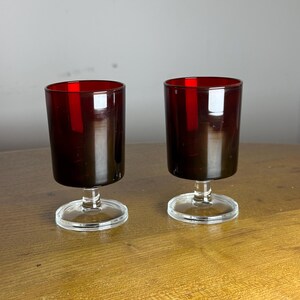 May include: Two stemmed glasses with a dark red colour. The upper part of the glasses is a deep red, fading to a darker shade towards the base. The stems and bases are clear glass, contrasting with the coloured bowls.