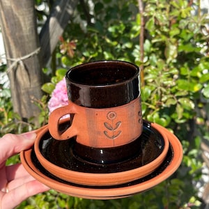 May include: A handcrafted ceramic coffee cup and saucer set. The cup is dark brown with a terracotta band featuring a floral design. The saucer is a matching dark brown colour with a terracotta rim. Ideal for enjoying coffee or tea.