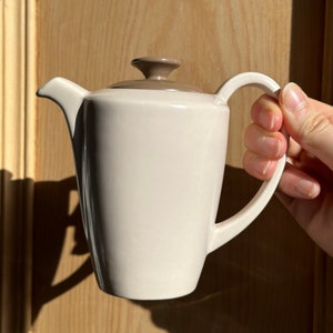May include: A cream-coloured ceramic teapot with a brown lid and handle. The teapot has a classic design with a curved spout and a rounded body. The handle is arched, and the lid has a small knob. The teapot is held by a person.