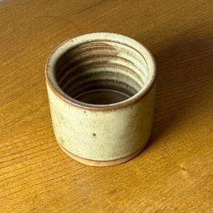 May include: A small, cylindrical ceramic container with a light green exterior and a brown interior. The container has a slightly textured surface and a rounded base. The interior features concentric brown rings. The container sits on a wooden surface.