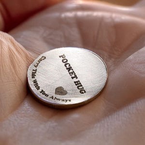 May include: A silver-colored, round pocket hug coin. The coin has the words "Pocket Hug" diagonally across the center, with "Carry This With You Always" around the edge. A small heart is etched below the words "Pocket Hug".