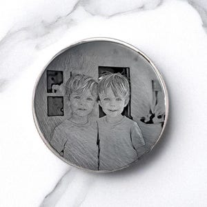 May include: A round, silver-toned pendant featuring a grayscale image of two young children. The image is etched into the pendant's surface, creating a unique, personalized keepsake. The pendant has a polished, reflective border.
