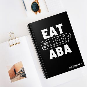Könnte beinhalten: Schwarzes spiralgebundenes Notizbuch mit dem Text "EAT SLEEP ABA" in Weiß. Das Notizbuch hat ein weißes Logo mit dem Schriftzug "Let's Study ABA" unten.
