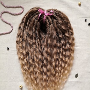 Peut inclure: Un paquet de longues extensions de cheveux bouclés avec un dégradé de couleur brun clair à blond. Les cheveux sont attachés ensemble avec un ruban rose.