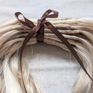 May include: Close-up of a bundle of blonde hair extensions tied with a brown ribbon. The hair is layered and appears to be natural in color. The ribbon is tied in a bow, adding a decorative element to the hair extensions.