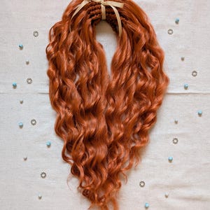 May include: A set of vibrant, copper-coloured curly hair extensions, tied with a light gold ribbon. The extensions are arranged in a heart shape, with a soft, wavy texture. Scattered around are small silver-coloured charms and light blue beads.