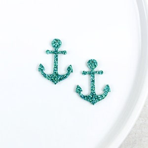 May include: Two teal glitter anchor charms. The anchors are made of a sparkly material and have a hole at the top for attaching to a necklace or bracelet.