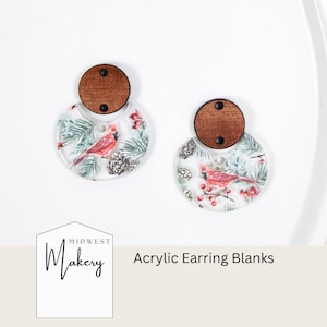 Puede incluir: Dos plantillas de pendientes de acrílico. Cada una tiene una parte superior de madera y una base circular transparente con un diseño invernal. La base presenta un cardenal rojo, agujas de pino y piñas. El texto "Acrylic Earring Blanks" es visible.