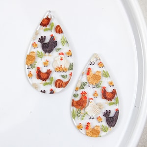 Puede incluir: Dos pendientes de acrílico transparente en forma de lágrima con un patrón colorido de pollos y pollitos.