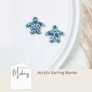 May include: Two blue and green acrylic turtle earring blanks on a white plate. The plate is on a white surface. The text "Midwest Makery" and "Acrylic Earring Blanks" is visible below the plate.