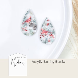 Puede incluir: Dos blancos de pendientes de acrílico transparente en forma de lágrima con un diseño invernal de cardenal y piña. Los blancos están sobre una superficie blanca. Blancos de pendientes de acrílico Midwest Makery.