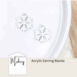 May include: Two white snowflake-shaped acrylic earring blanks. The blanks are on a white surface. The text "Midwest Makery Acrylic Earring Blanks" is visible below the blanks.