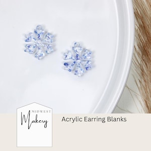 May include: Two snowflake-shaped acrylic earring blanks with a blue and white glitter design. The blanks are on a white plate. The text "Midwest Makery" and "Acrylic Earring Blanks" is visible in the image.