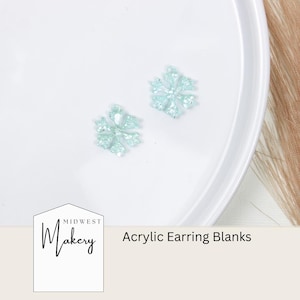 May include: Two light blue snowflake-shaped acrylic earring blanks on a white plate. The plate is on a light brown surface. The text "Midwest Makery" and "Acrylic Earring Blanks" is visible below the plate.