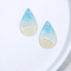 May include: Two teardrop-shaped resin earrings with a beach scene design. The earrings feature a blue ocean with white waves crashing on a sandy beach.