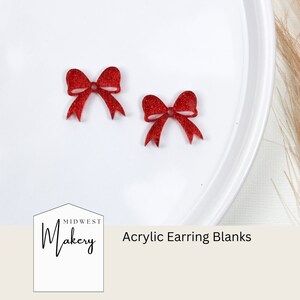 May include: Two red glitter bow acrylic earring blanks on a white surface. The text "Midwest Makery" and "Acrylic Earring Blanks" is visible below the bows.