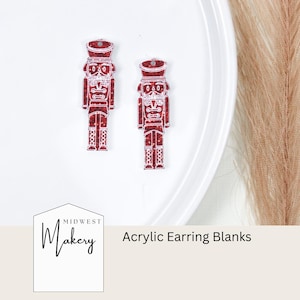 May include: Two red glitter nutcracker acrylic earring blanks with white details. The blanks are on a white plate. The text "Midwest Makery" and "Acrylic Earring Blanks" are visible below the blanks.