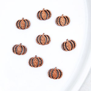 Pode incluir: Sete pequenos ornamentos planos em forma de abóbora dispostos de forma dispersa. Cada abóbora apresenta um acabamento texturizado com glitter na cor cobre na superfície frontal com linhas verticais escuras recortadas definindo os segmentos.