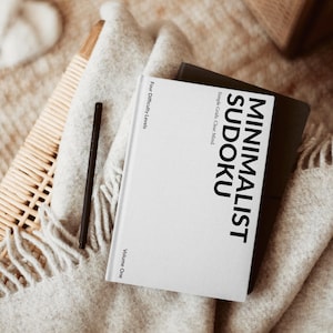 Op de afbeelding: Een close-up van een "Minimalist Sudoku" boek met zwarte tekst en "Volume One" zichtbaar. Een zwarte pen ligt op een lichtgrijze deken. De scène speelt zich af op een geweven oppervlak met een houten doos op de achtergrond.