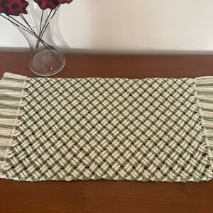 May include: A rectangular macrame table runner with a woven diamond pattern in off-white and green. The ends feature striped fringe in matching colors. The runner is displayed on a wooden surface.