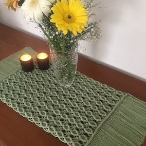 May include: A green macrame table runner with fringe detail, displayed on a wooden surface. A clear glass vase holds a bouquet of yellow and white gerbera daisies and baby's breath. Two lit candles in decorative holders are on the runner.