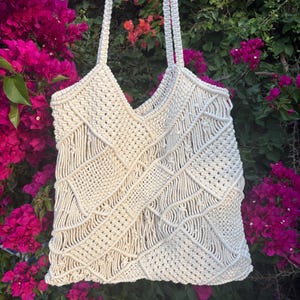 Peut inclure: Sac fourre-tout en macramé crème avec un motif géométrique. Le sac présente une ouverture encolure en V et de longues anses tressées. Il est suspendu devant un fond de fleurs roses et oranges vives.