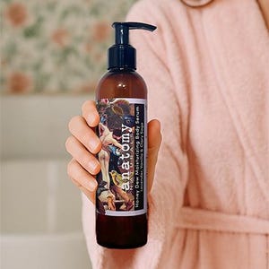 Honey Dew botanical body serum held in a woman’s hand with bathrobe visible, intensive post shower body serum for dry and sensitive skin.